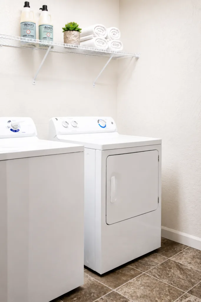 A clean laundry room with a white washer and dryer set on a tiled floor, shelves above holding detergent and rolled towels.