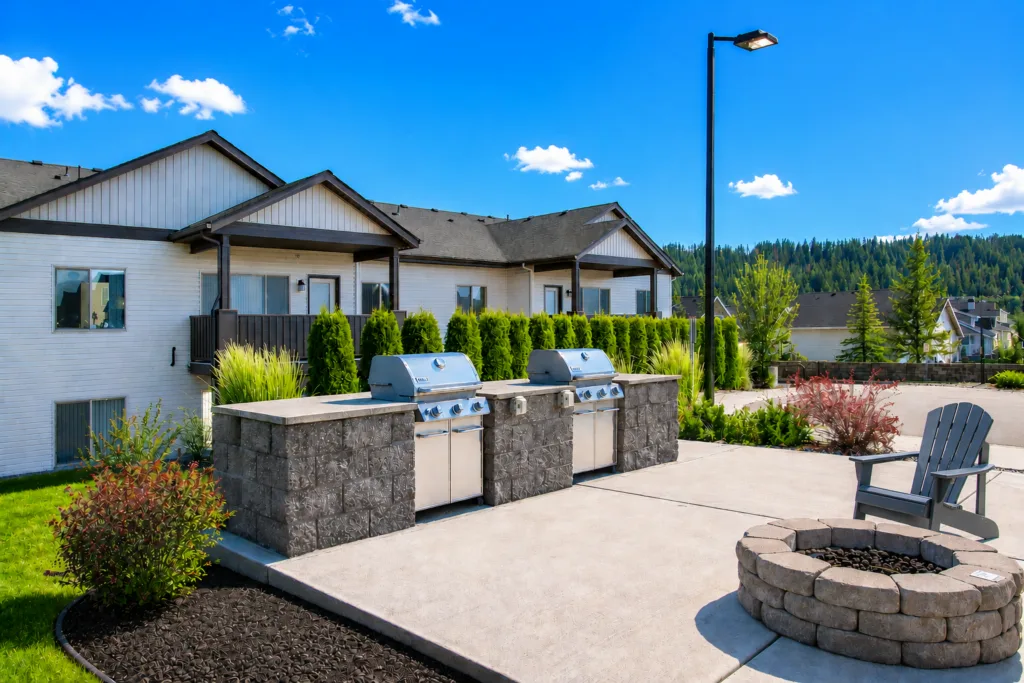 A patio with two stainless steel grills, a fire pit surrounded by chairs, and residential buildings in the background.
