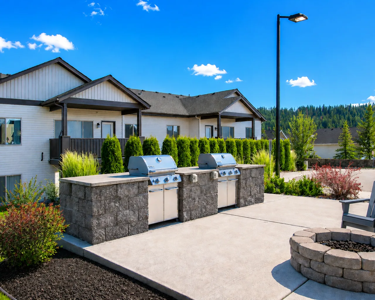 Outdoor area with two stainless steel grills on a stone platform, surrounded by greenery and a residential building in the background.