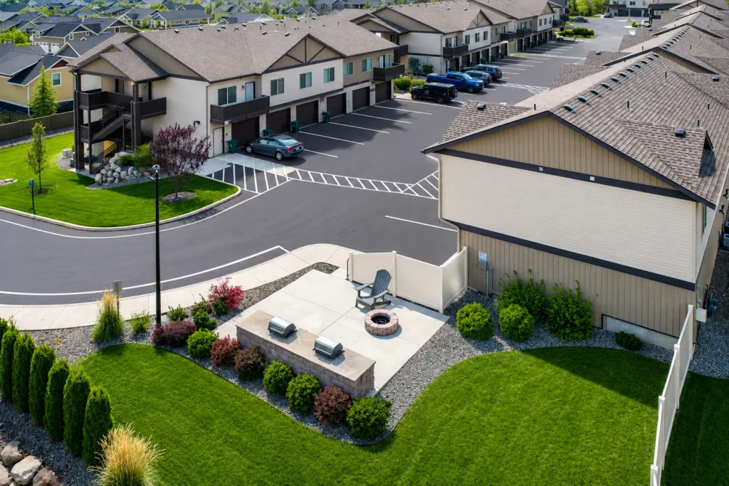 Aerial view of a residential area featuring apartment buildings, a parking lot with cars, and a landscaped common area with benches and a fire pit.