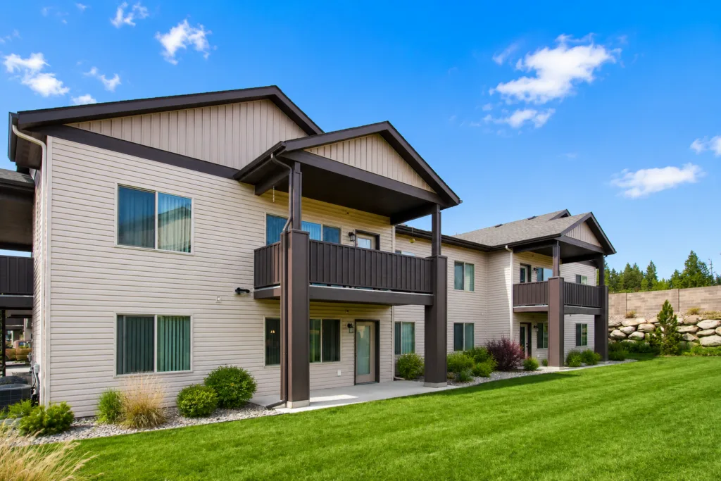 A modern two-story apartment building with balconies, surrounded by a neatly maintained lawn under a clear blue sky.