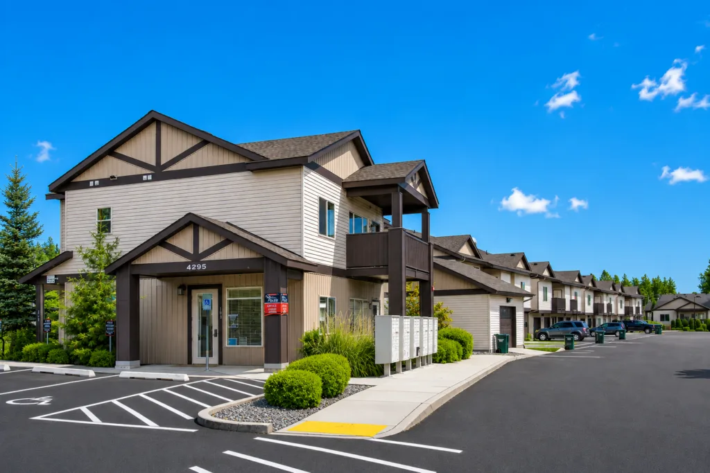 Apartment complex with beige siding and dark trim, featuring a parking lot and clear blue sky.