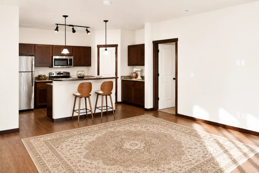 A modern kitchen and dining area with wood cabinets, a stainless steel refrigerator, bar stools at a counter, and a patterned rug.
